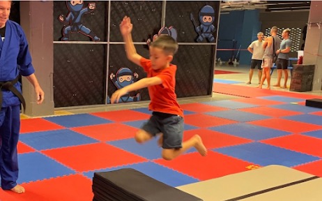 A boy mid-air over the mats at NinjaGym while Rick watches